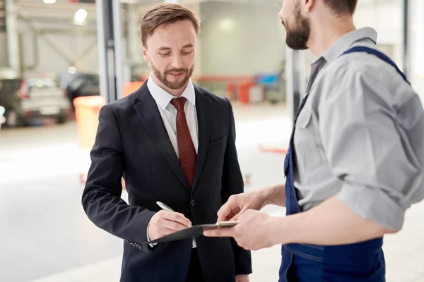 Waist up portrait of handsome businessman signing contract for car repairs while talking to bearded mechanic in service garage