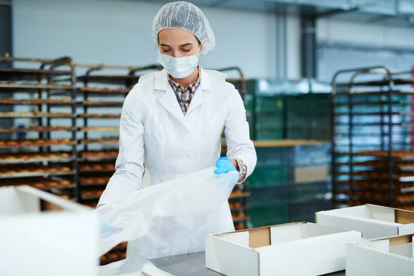 Confectionery factory employee in white coat holding baking parchment to put into empty package.