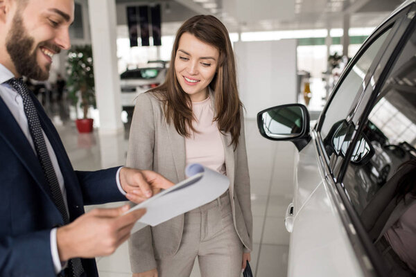 Portrait of beautiful young woman smiling happily while listening to car dealer  choosing luxury car in showroom