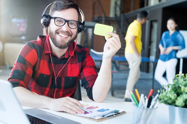 Portrait of smiling bearded man wearing headphones and glasses showing blank green card while sitting at desk in modern office of IT developers company, copy space
