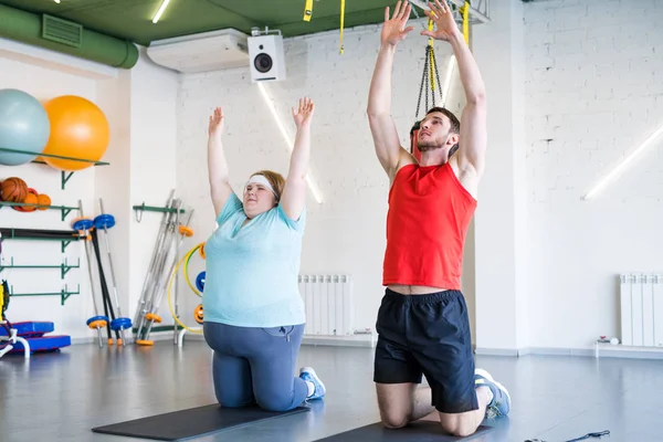 Full length portrait of obese young woman doing fitness exercises in gym with personal trainer helping her