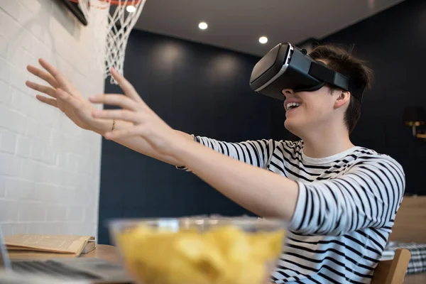 Excited young male extending hands forward while using virtual reality headset sitting at home.