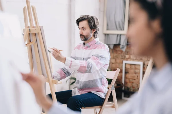 とした日当たりの良いロフト スタジオで仕事を楽しんでいる絵画美術の授業でイーゼルに座っている中年の男性像側ビュー — ストック写真
