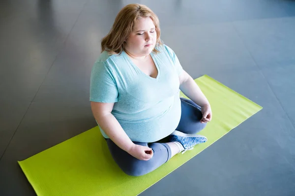 High angle portrait of obese young woman sitting in lotus position and meditating with eyes closed