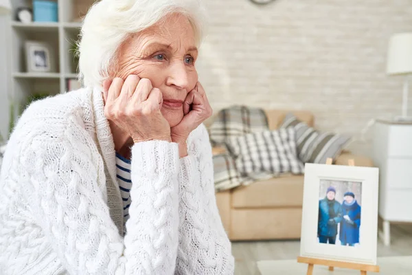 Portrait of lonely senior woman staring into space with tears in her eyes, sitting by picture of  her family, copy space (photo in frame by me)