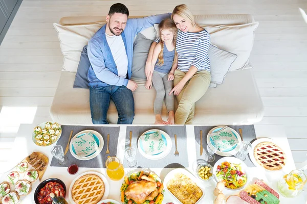 High angle portrait of successful family enjoying dinner together in modern apartment sitting on couch with adorable little girl, all smiling happily