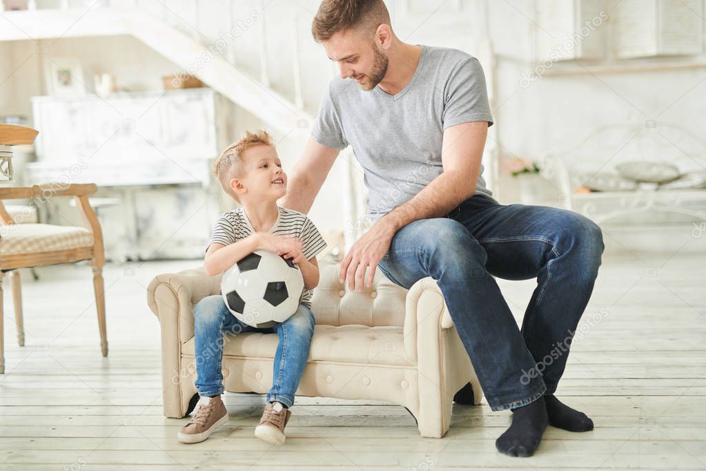 Full length warm toned  portrait of caring father talking to cute little son holding football ball sitting on small couch at home