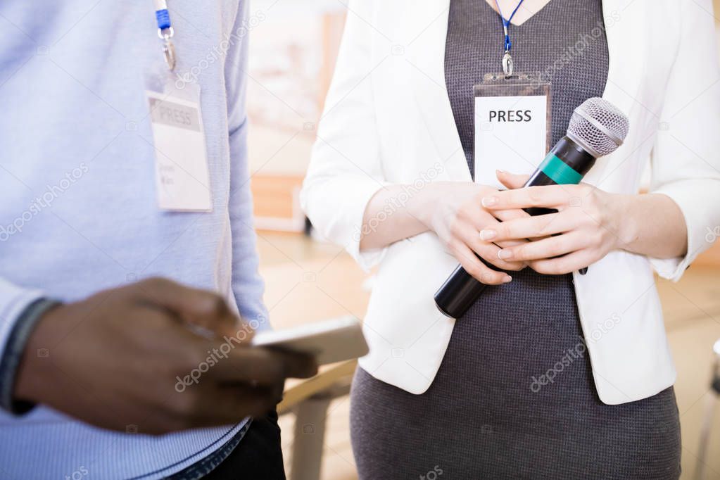 Mid section closeup of two journalists holding microphones standing at press conference with ID tags in focus