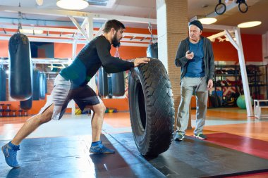 Saygısız ve ağır lastik eğitim boks jimnastik salonu profesyonel koç ile iterek kendine güvenen sporcu yan görünüm.