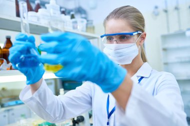 Laboratuarda çalışırken şişeler sıvılarda çözümlenirken rengini ciddi düşünceli genç kadın bilim Ameliyat maskesi ve koruyucu gözlükler