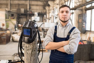 Ciddi emin sakallı genç yüksek profesyonel kaynakçı ile çapraz silah gaz silindir maskesi kaynak ve kamera fabrika dükkan bakarak ile ayakta