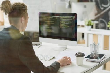 Rear view of hipster coder with hair bun wearing jacket using computer while programming app and sitting at desk with technologies and coffee cup clipart