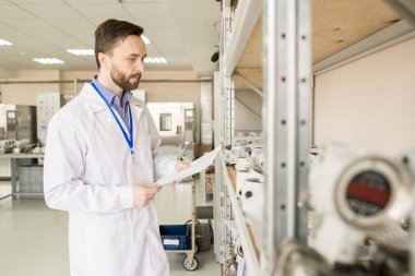 Orta yaşlı fabrika işçisi ölçüm cihazları elinde manometreler, iç arka plan üzerinde geniş ambarının doğru işlem denetlerken listesini tutan önlük giyen portre çekim