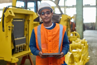 Bel öğe, dikey, yansıtıcı yelek ve kask elinde, arka plan üzerinde üretim bölümünün iç dijital tablet tutarken kameraya bakarak giymiş yakışıklı karma yarış makinesi operatörü