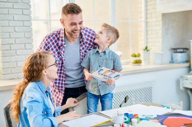 Sandalye üzerinde duran ve verirken ona el yapımı tebrik kartı babalar günü, modern oturma odası arka plan üzerinde iç için geniş bir gülümseme ile gülen babası seyir sarı saçlı küçük çocuk