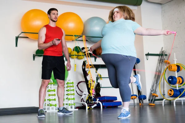 Full length portrait of obese woman jumping with rope in gym while training with fitness instructor