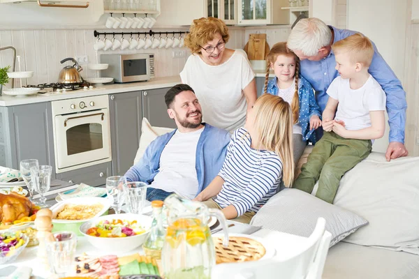 Portrait of happy two generation family enjoying dinner party together in modern apartment standing round couch with two adorable children in sunlight