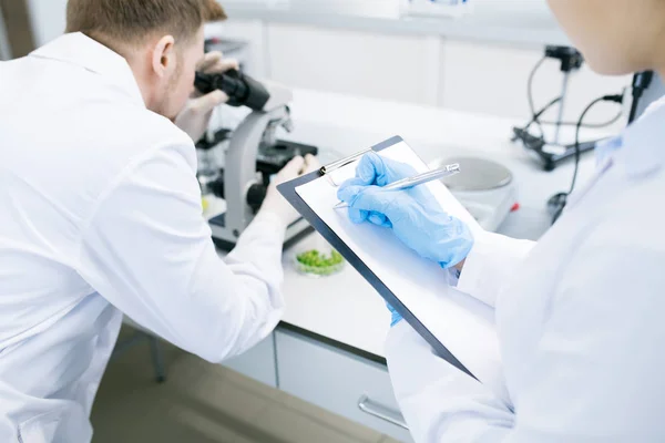 裁剪后视图女性微生物学家写下来到剪贴板中从人收到的数据分析绿色蔬菜结构在显微镜下的研究 — 图库照片