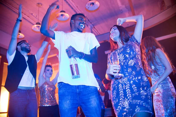 Low angle view of cheerful friends standing in circle at modern night club and dancing with raised hands, multi-ethnic couple with alcoholic drinks on foreground