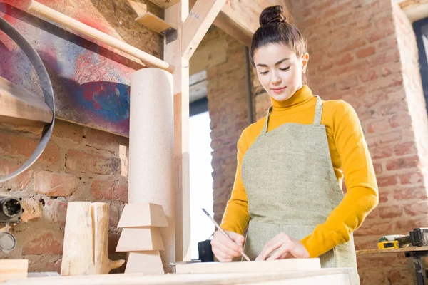 Waist up portrait of pretty young woman wearing apron working with wood in crafting shop, standing against brick wall