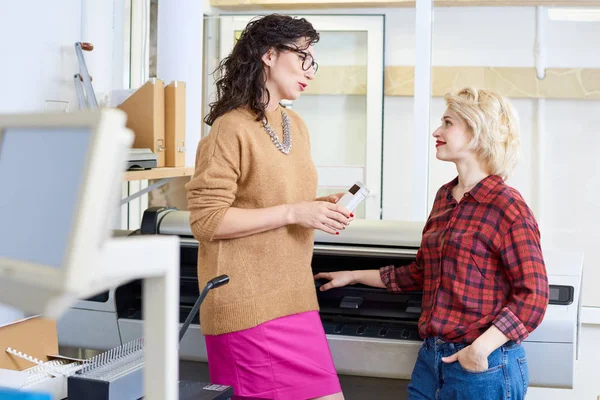 Two female printing operators standing near plotter in office and having nice conversation.