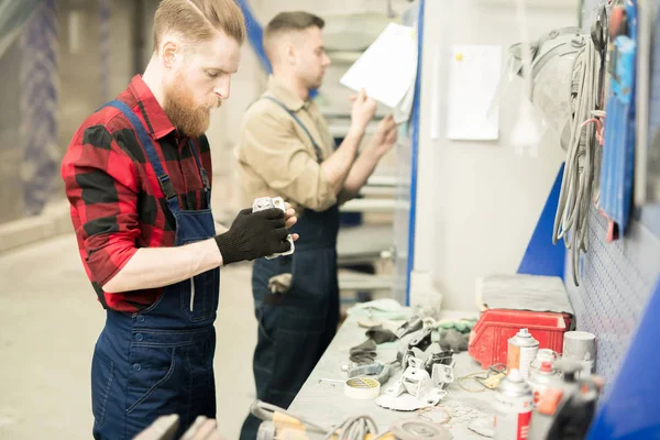 Young bearded male automotive technician selecting necessary spare parts while his colleague checking schedule in service garage