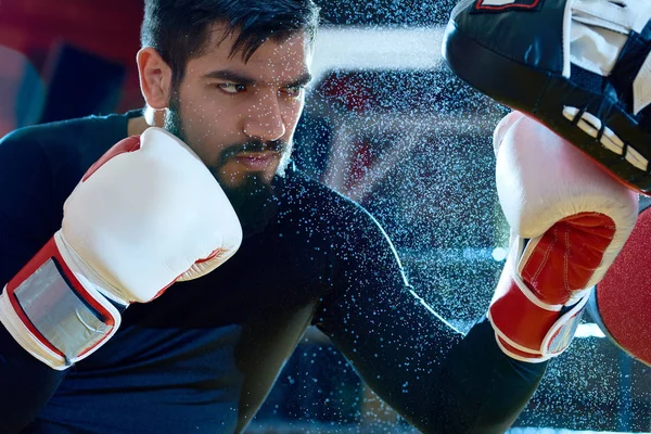 Bearded determined man boxing on ring with trainer while punching focus mitts in water drops.