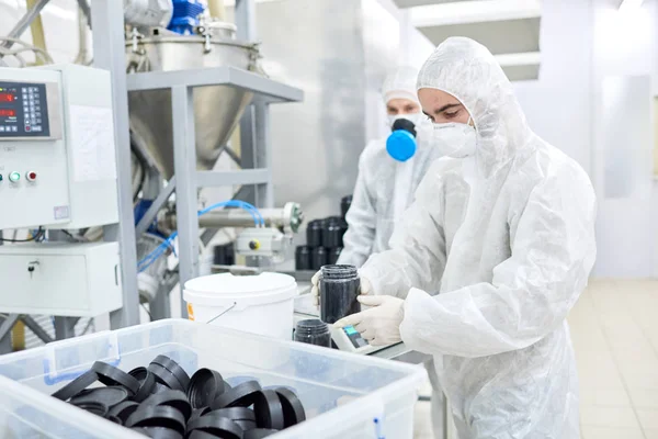 Production worker in protective clothing carefully carrying open jar with sports nutrition.