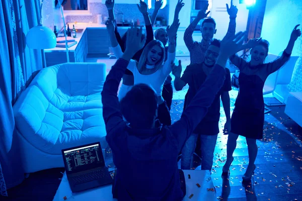 Group of modern young people dancing with hands in air  listening to DJ playing music at private house party, lit by blue light