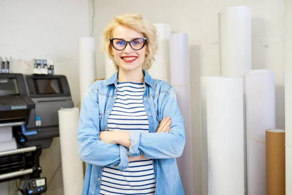 Lovely young woman in stylish outfit smiling and looking at camera while standing in middle of printing office.