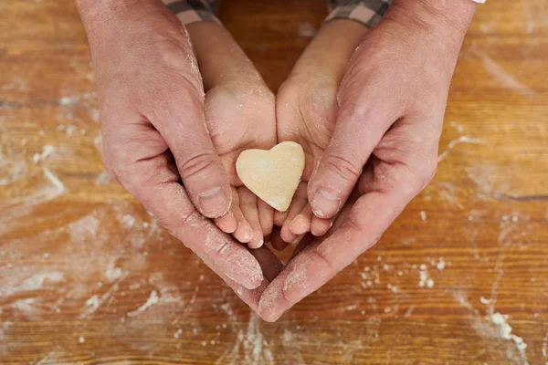 Hands of father and his little daughter holding unprepared heart-shaped cookie together, wooden table covered with flour on background