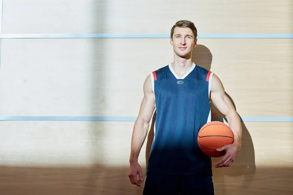 Handsome man in basketball uniform smiling and looking at camera while holding ball and standing near gym wall.
