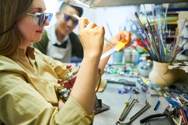 stock image Side view portrait of young female artist melting glass by gas torch while making beads for beautiful handmade jewelry in glassworking studio
