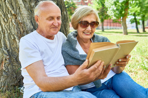Portrait of modern senior couple sitting under tree in park enjoying sunny summer day and reading book