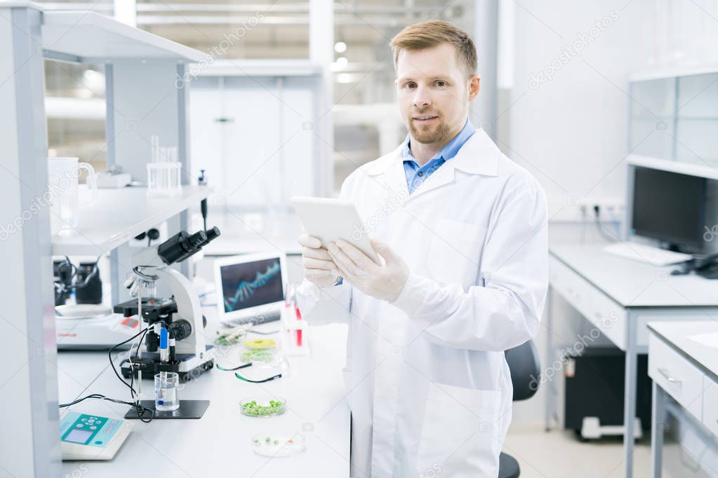 Young male microbiologist in laboratory coat standing at desk with microscope and green vegetable samples and holding tablet smiling and looking at camera