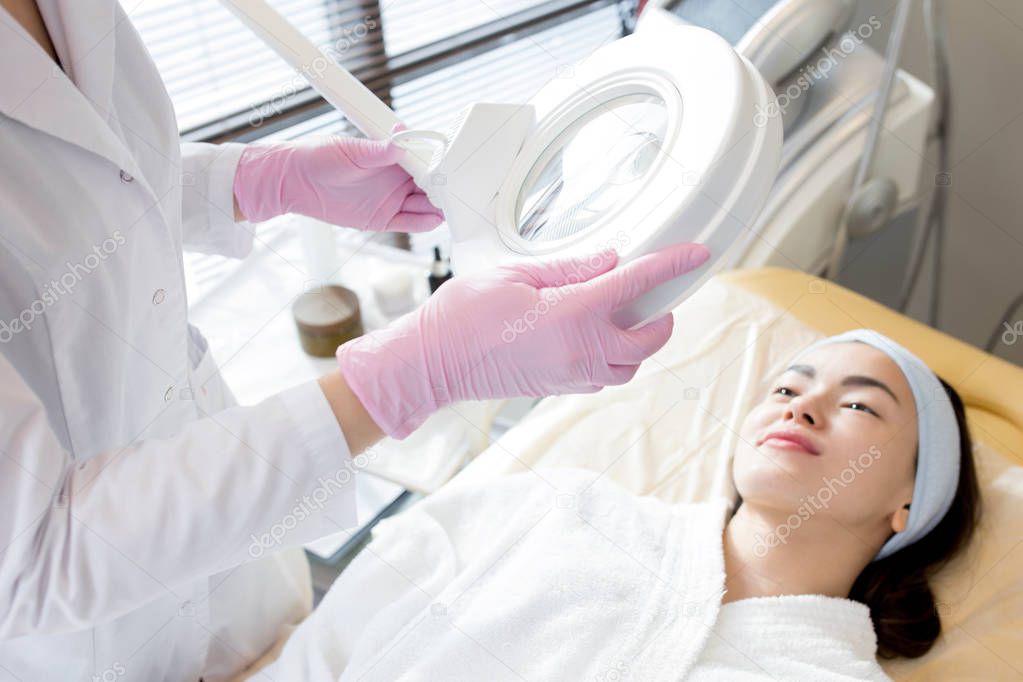 Unrecognizable beautician wearing white coat and rubber gloves using magnifying lamp while examining facial skin of pretty young client lying on treatment table