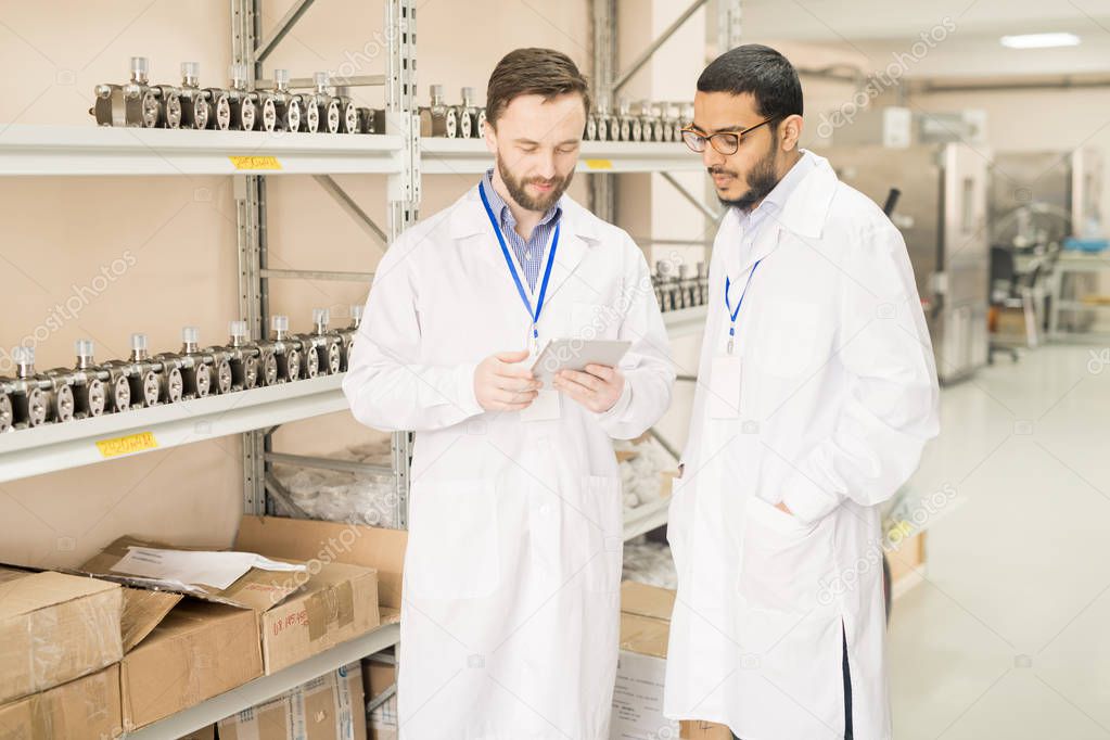 Highly professional team of measuring equipment factory workers wearing lab coats gathered together at spacious warehouse and analyzing results of accomplished work with help of digital tablet.