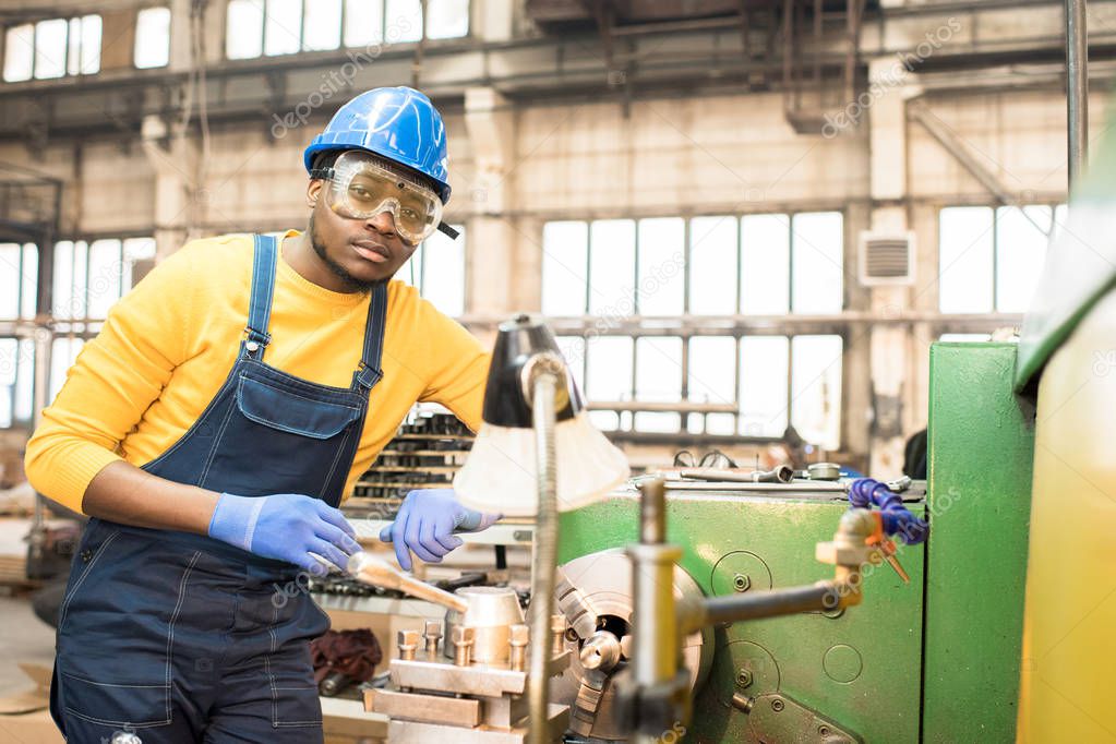 Portrait shot of handsome young lathe operator wearing protective helmet and safety goggles standing at spacious production department of modern plant and looking at camera