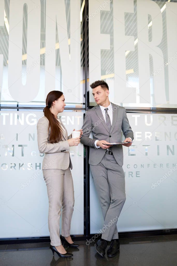 Full length portrait of two business people, man and woman, talking to each other at coffee break  standing in hall of modern office building