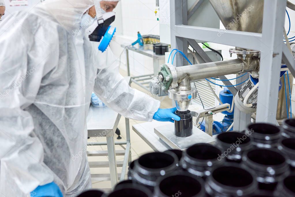 Production worker standing in protective clothing and filling black jar with sports nutrition.