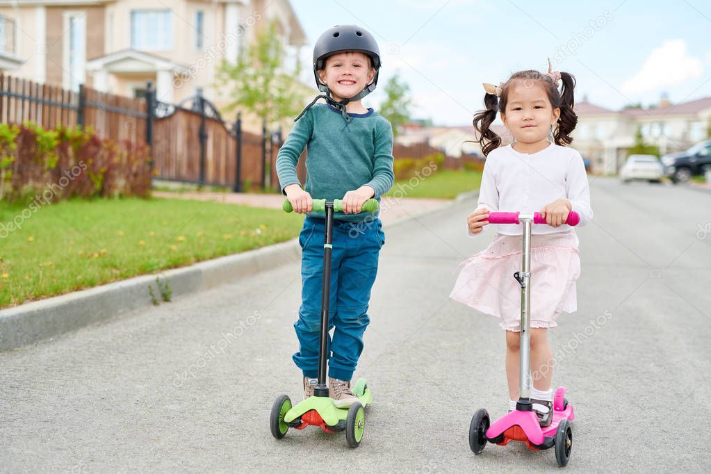 Full length portrait of two cute children, boy and girl,  riding three wheel scooters and smiling happily while going down road in rich residential area, copy space
