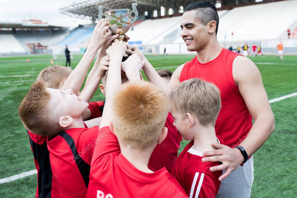 Portrait of junior football team holding trophy together and cheering after winning match in outdoor stadium