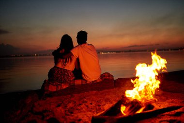 Kamp ateşi arkalarındaki geceleri yanan plajda, sakin deniz düşündüğünü süre ekose ve kucaklayan arkadan görünüş romantik çift kütük üstünde oturan kaplı