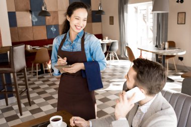 Neşeli genç ve güzel karışık yarış garson önlük müşterinin o üstünde hareket eden telefon modern restoranda öğle yemeği sırasında söz ederken emir alan