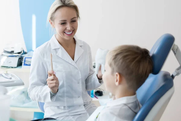 歯科用椅子に座っている少年に口腔衛生ルールを説明する歯ブラシと歯の模型を持って笑顔の女性歯科医の肖像画 — ストック写真