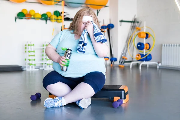 Full length portrait of exhausted obese woman wiping sweat with towel sitting on step after fitness training