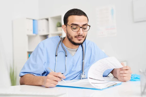 Portrait of young Middle-Eastern doctor wearing glasses sitting at desk in office and reading patients reports