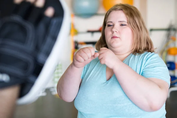 Waist up portrait of obese young woman working out in gym with personal fitness coach, hitting pads