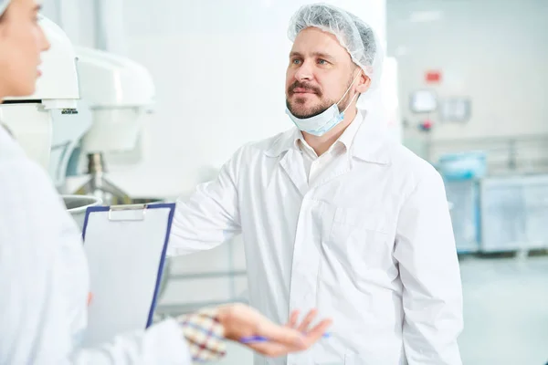 Confectionery factory employee standing in white coat and discussing with colleague holding clipboard.