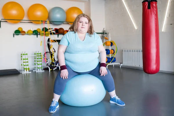 Full length portrait of young obese woman sitting on fitness ball looking at camera